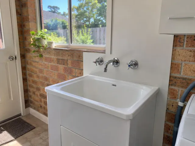 White laundry sink with new chrome tap under a window and brick wall in Forestville